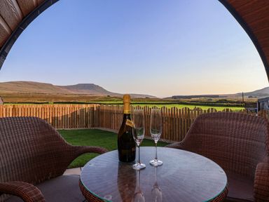 A patio with two wicker chairs around a glass table with a bottle of sparkling wine and two wine glasses overlooking fields and hills at Curlew in Chapel Le Dale near Ingleton