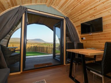Interior of a wooden pod with a dining table and chairs next to double glass doors opening to a fenced yard and distant hills at Curlew in Chapel Le Dale near Ingleton
