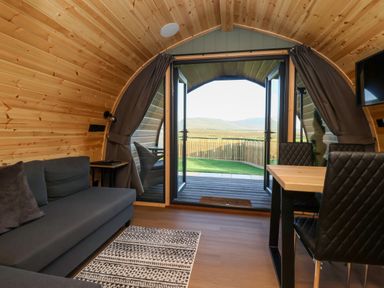An interior view of a wooden pod with an open double door showing a fenced yard and hills beyond a gray sofa a dining table with black chairs and a rug on wooden floor Curlew Chapel Le Dale near Ingleton