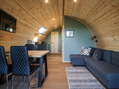 An interior of a wooden curved room with a black TV on the wall a dining table with six black chairs a gray sectional sofa with cushions and a patterned rug at Curlew in Chapel Le Dale near Ingleton