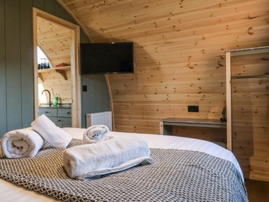 A bedroom with a bed holding rolled towels a television mounted on a wall and a kitchenette visible through a doorway at Curlew in Chapel Le Dale near Ingleton