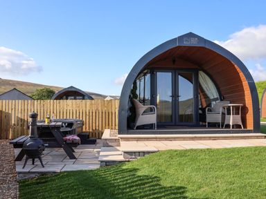 An outdoor view of a wooden pod with wicker chairs on a patio and a picnic table with a chiminea at Curlew in Chapel Le Dale near Ingleton