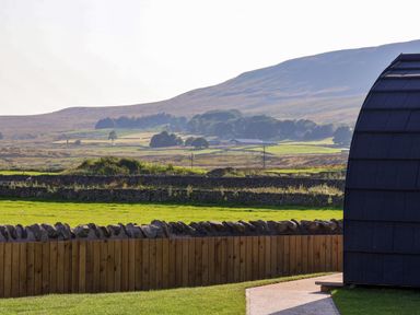 A grassy yard with a wooden fence and a curved roof structure with hills in the background at Curlew in Chapel Le Dale near Ingleton