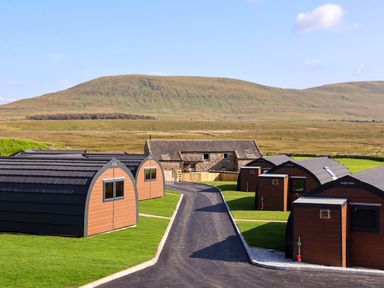 A group of wooden pod cabins along a paved path with grassy areas and a barn in the background at Curlew in Chapel Le Dale near Ingleton