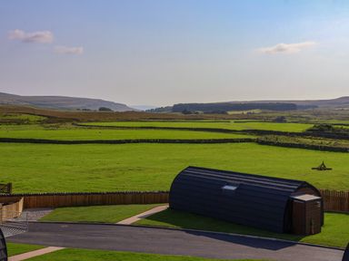 A green field with a dark curved shed and a paved path in a rural area at Woodpecker in Chapel Le Dale near Ingleton