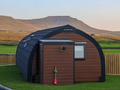 A small curved wooden cabin with a window and a sign that reads woodpecker in a grassy area with mountains in the background at Woodpecker in Chapel Le Dale near Ingleton