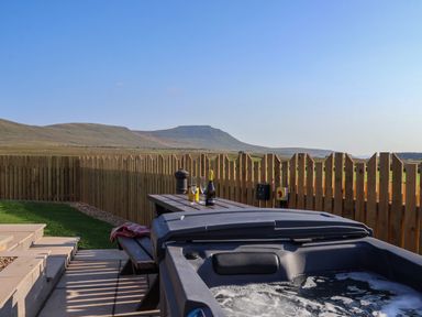 A backyard with a bubbling hot tub a picnic table with glasses and a bottle and a wooden fence with hills in the background at Woodpecker in Chapel Le Dale near Ingleton