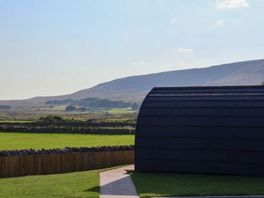 A curved roof building with a stone wall and green fields with hills in the background at Woodpecker in Chapel Le Dale near Ingleton