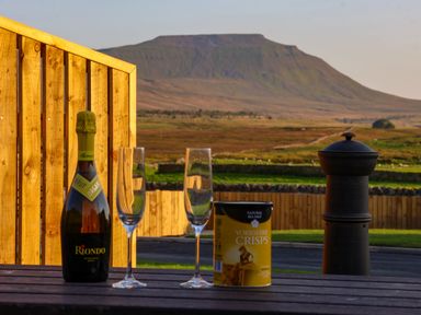 A bottle of sparkling wine two empty glasses and a can of Yorkshire crisps on a wooden table outside with a mountain and green fields in the background at Woodpecker in Chapel Le Dale near Ingleton