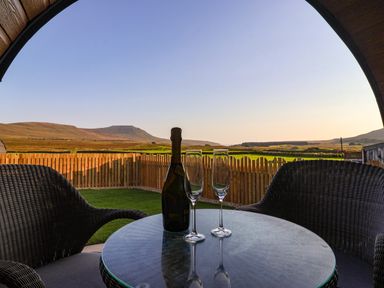 A round glass table with a bottle and two empty champagne glasses between two wicker chairs overlooking a fenced grassy area and distant hills at Woodpecker in Chapel Le Dale near Ingleton