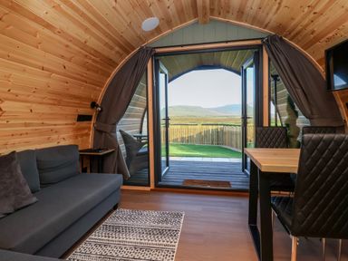 A wooden interior room with a gray sofa a table with black chairs and an open door showing a fenced yard and hills at Woodpecker in Chapel Le Dale near Ingleton