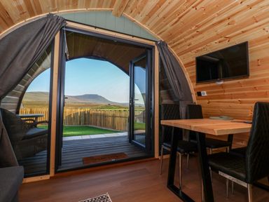 A wooden pod interior with a dining table and chairs and an open door showing a fenced yard and hills at Woodpecker in Chapel Le Dale near Ingleton