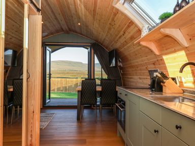 A kitchenette with appliances and dining table with chairs near open doors showing a fenced yard and hills at Woodpecker in Chapel Le Dale near Ingleton