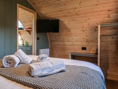 A bedroom with rolled towels on a bed a wall mounted tv a small kitchenette visible through a doorway and wooden walls at Woodpecker in Chapel Le Dale near Ingleton