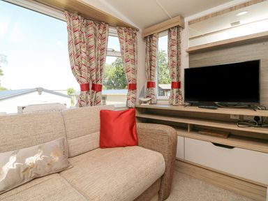 A seating area with a beige sofa and cushions near windows with patterned curtains and a TV on a wooden stand at 25 Manleigh Park Combe Martin