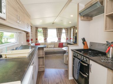 Kitchen area with stove sink and cabinets leading to a living room with sofa and windows at 25 Manleigh Park in Combe Martin