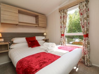 A bedroom with a bed with white and red bedding next to a window with patterned curtains at 25 Manleigh Park Combe Martin