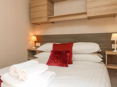 A bedroom with a bed with white linens red pillows folded towels on the bed and bedside lamps at 25 Manleigh Park in Combe Martin