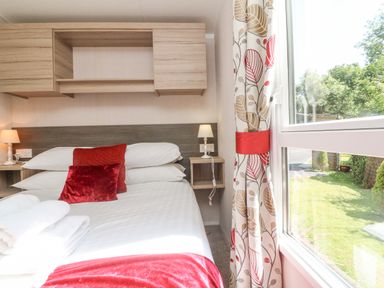 A bedroom with a bed with red pillows and white sheets next to a window with patterned curtains at 25 Manleigh Park in Combe Martin
