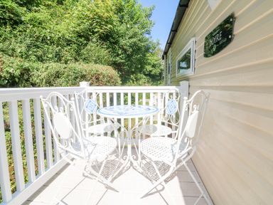 A small outdoor balcony with white metal table and chairs surrounded by white railing and greenery at 25 Manleigh Park in Combe Martin