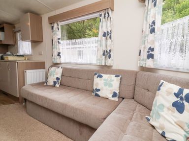 A living area with a beige L-shaped sofa with floral cushions and patterned curtains at 26 Manleigh Park in Combe Martin