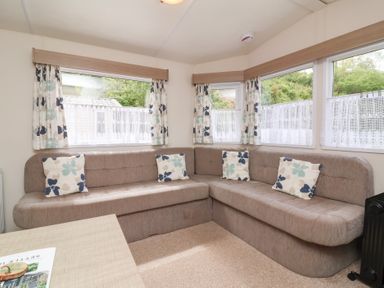 A corner seating area with a brown upholstered sofa and floral cushions by windows with curtains at 26 Manleigh Park in Combe Martin