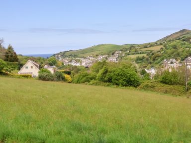 A grassy field with bushes and houses in the background with hills and a distant view of the sea at 26 Manleigh Park in Combe Martin