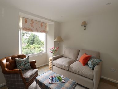 A living room with a beige sofa and an armchair next to a window with floral curtains at Little Robin Cottage in Ambleside