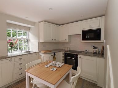A kitchen with white cabinets a wooden table with two chairs and a window with flowers at Little Robin Cottage in Ambleside