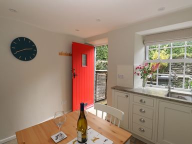 A kitchen with a wooden table set with a bottle and two glasses near a window with flowers and an open red door at Little Robin Cottage in Ambleside