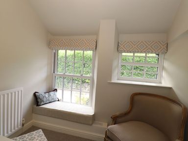 A corner of a room with two windows one having a cushioned window seat and a patterned cushion and the other with a beige upholstered armchair at Little Robin Cottage in Ambleside