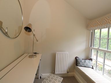 A corner with a white desk, metal lamp, round wall mirror, radiator, window seat with cushion and outside greenery at Little Robin Cottage in Ambleside