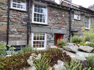 A stone cottage exterior with white framed windows plants and rocks near the entrance at Little Robin Cottage in Ambleside
