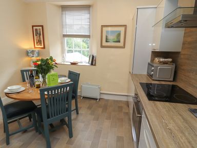 A kitchen with a round table set for four with chairs and flowers on the table a window a microwave and a stovetop at Belmont House in Bowness-On-Windermere