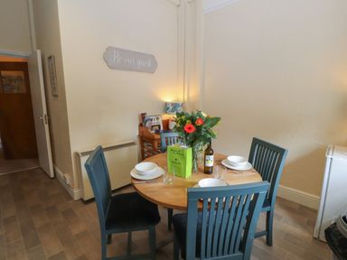 A dining area with a round wooden table set for four with bowls and glasses with blue chairs and a flower vase on the table at Belmont House in Bowness-On-Windermere