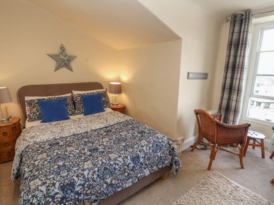 A bedroom with a bed with floral bedding two bedside tables with lamps a wicker chair by a window with plaid curtains at Belmont House in Bowness-On-Windermere