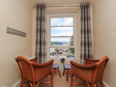 A small seating area with two wicker chairs and two stools by a window with plaid curtains at Belmont House in Bowness-On-Windermere