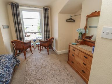 A small room with two wicker chairs and tables by the window a wooden dresser with a mirror and a rug on the floor at Belmont House in Bowness-On-Windermere