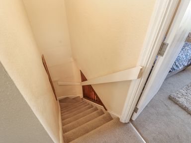 A narrow carpeted staircase with a wooden handrail and walls leading downstairs next to a doorway showing part of a bedroom at Belmont House in Bowness-On-Windermere