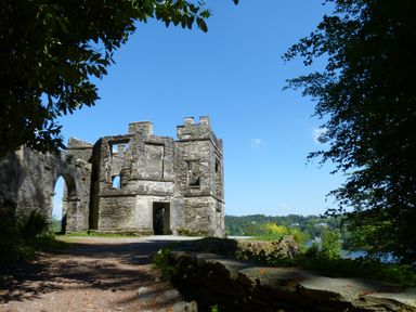 Stone ruins of a building with archways on a path surrounded by trees and overlooking a distant landscape with water