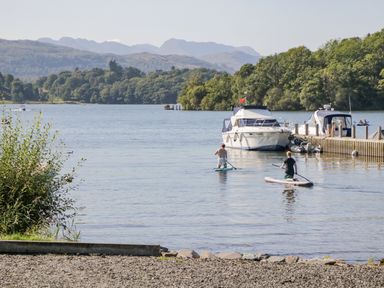 Two people paddleboarding on a lake near boats docked at a pier surrounded by trees and hills at Belmont House in Bowness-On-Windermere