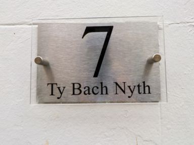 A metal house number sign with the number 7 and the text Ty Bach Nyth on a white wall at Hole in the Wall in Caernarfon