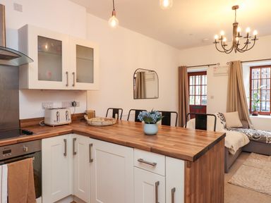 A kitchen with wooden countertops and white cabinets next to a dining area with a table and chairs and a living area with a sofa and windows at Hole in the Wall in Caernarfon