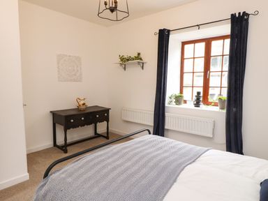 A bedroom with a bed near a window with curtains and a wooden table with a decorative pitcher at Hole in the Wall in Caernarfon