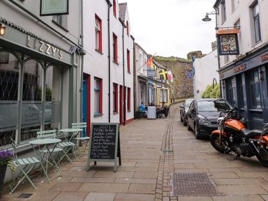 A narrow street with outdoor seating at Lizzy's and Four Alls pub with parked cars and a motorcycle at Hole in the Wall in Caernarfon