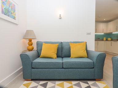 A blue sofa with two yellow cushions a yellow lamp on a side table and a geometric rug in a living room with a kitchen in the background at Rolles Rainbow in Exmouth