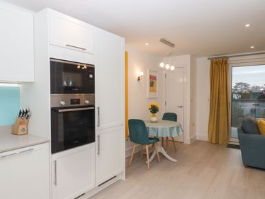 A kitchen area with built in ovens and a small dining table with two chairs and a vase of flowers next to a living room with yellow curtains at Rolles Rainbow in Exmouth