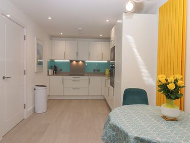 A kitchen with white cabinets blue glass backsplash and a table with a blue patterned tablecloth and yellow flowers in a vase at Rolles Rainbow in Exmouth