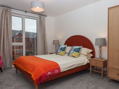 A bedroom with a double bed with an orange headboard and colorful pillows two bedside tables with lamps a wardrobe and large window with curtains at Rolles Rainbow in Exmouth