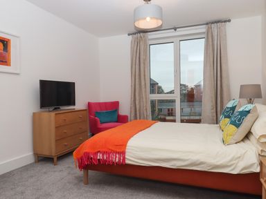 A bedroom with a bed an orange throw a wooden dresser a television a red armchair and a window with curtains at Rolles Rainbow in Exmouth